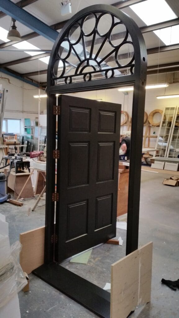 Black painted timber entrance door with traditional panel detailing, fitted with an elegant curved fanlight featuring decorative black metal glazing bars. The door is shown part-open in a joinery workshop during final quality checks.