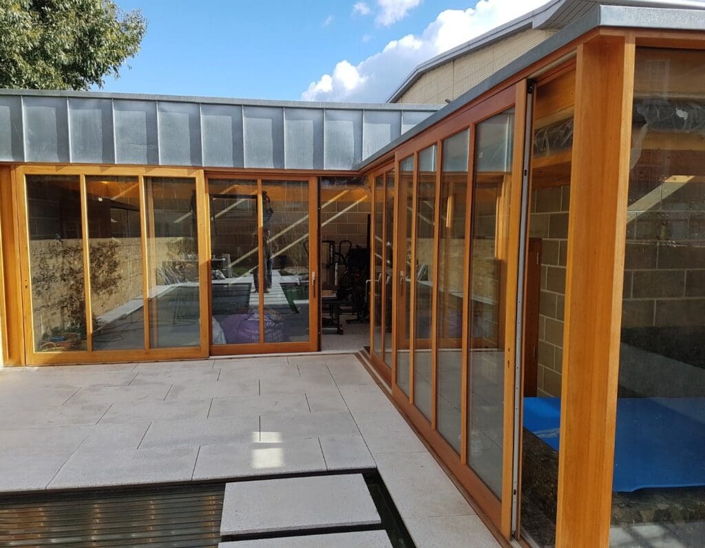 Timber lift and slide doors with a natural stained finish forming a corner configuration around a modern home extension. The large glass panels create a seamless view into the interior, which includes a gym and seating area, while reflecting the outdoor courtyard and blue sky above.