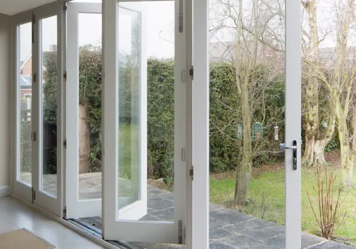 A white timber sliding-folding door system partially opened, revealing a garden with tall hedges and leafless trees in winter. The door panels fold neatly to the side, creating an open indoor-outdoor space.