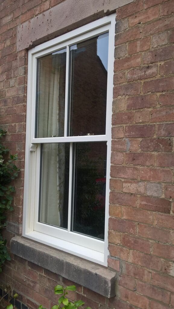 Close-up of a white-painted hardwood sliding sash window with Victorian-style astragal bars, set in a red brick wall with a stone lintel above.