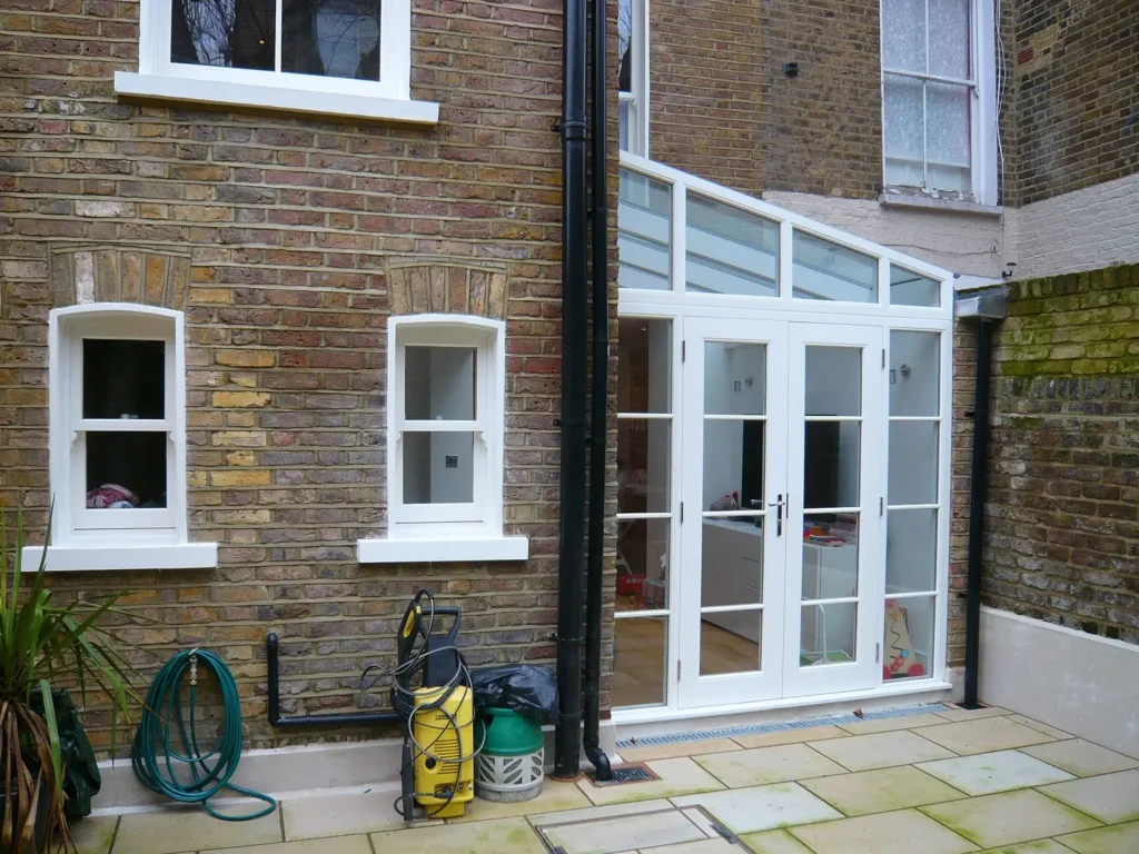 A rear view of a traditional London brick townhouse featuring bespoke white-painted Accoya sash windows and a matching glazed door set within a modern glass-roofed extension. The heritage-style windows blend seamlessly with the original architecture, while the clean lines and durability of Accoya timber highlight its suitability for bespoke restoration in period properties, as showcased in the Chesterton Road case study.