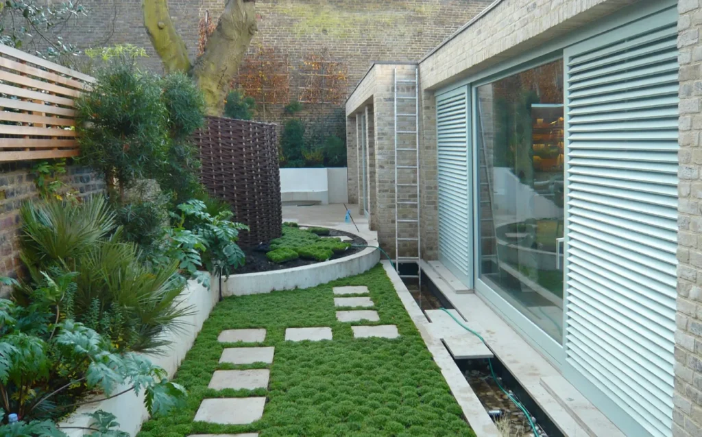 A sleek, contemporary courtyard garden with a modern brick-built home featuring large tilt-and-slide patio doors. The expansive glass panels are framed in soft green and paired with external louvred shutters, offering both aesthetic appeal and practical ventilation. The carefully landscaped garden path and lush planting create a tranquil urban retreat, demonstrating the stylish integration of Gowercroft’s tilt-and-slide door system in a high-end residential setting.