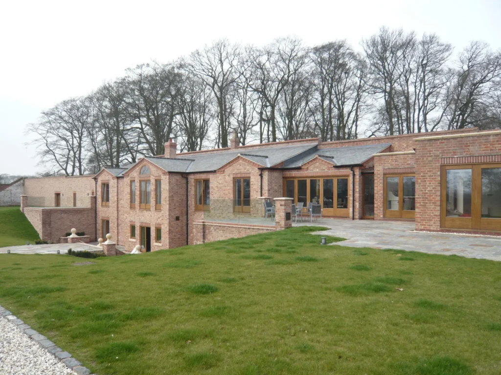 A large, elegant countryside home in Solihull constructed from traditional red brick, featuring extensive timber-framed windows and doors that blend harmoniously with the property’s classic architecture. The house overlooks a landscaped garden and patio area, showcasing Gowercroft’s premium timber glazing solutions, which bring warmth, character, and exceptional performance to this luxury residential development at Canwell Gardens.