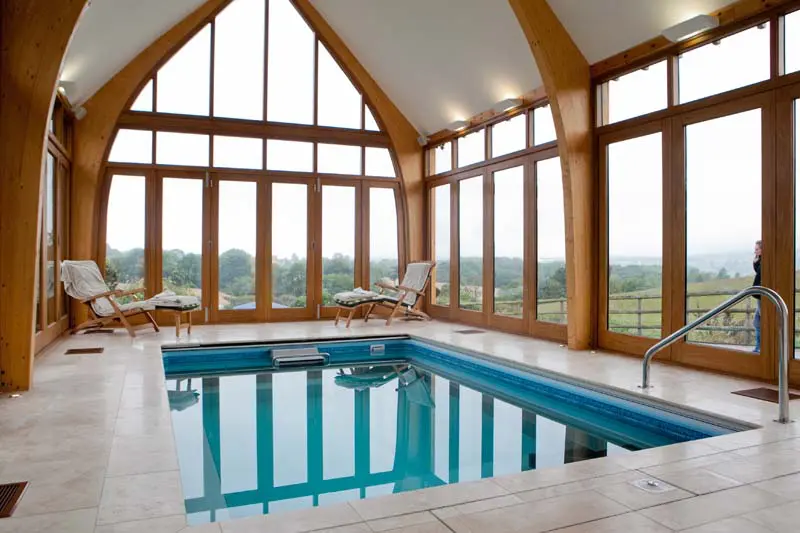 A luxurious indoor swimming pool enclosed by expansive timber bifold windows and doors, set within a vaulted oak-framed structure at Lumleigh House in Derbyshire. The glazing floods the space with natural light and offers uninterrupted views of the surrounding countryside, illustrating Gowercroft’s precision-crafted bifold solutions that combine elegance, functionality, and thermal performance in high-end residential settings.