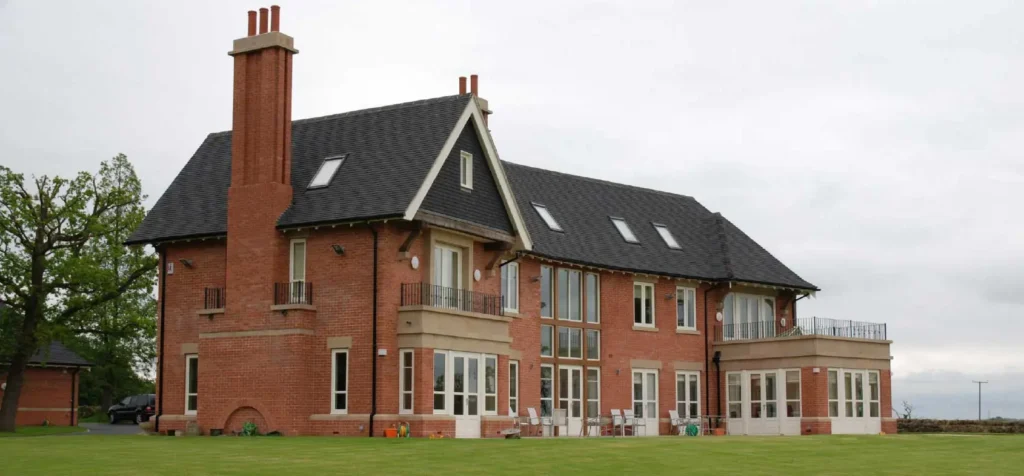 A grand, traditionally styled red-brick home set within a spacious landscaped garden, featuring multiple floors and distinctive architectural details such as tall chimneys, dormer windows, and Juliet balconies. The property is fitted with elegant white-painted timber windows and French doors, showcasing Gowercroft’s craftsmanship in providing bespoke wooden windows and doors for luxury residences like The Pastures in Derbyshire.