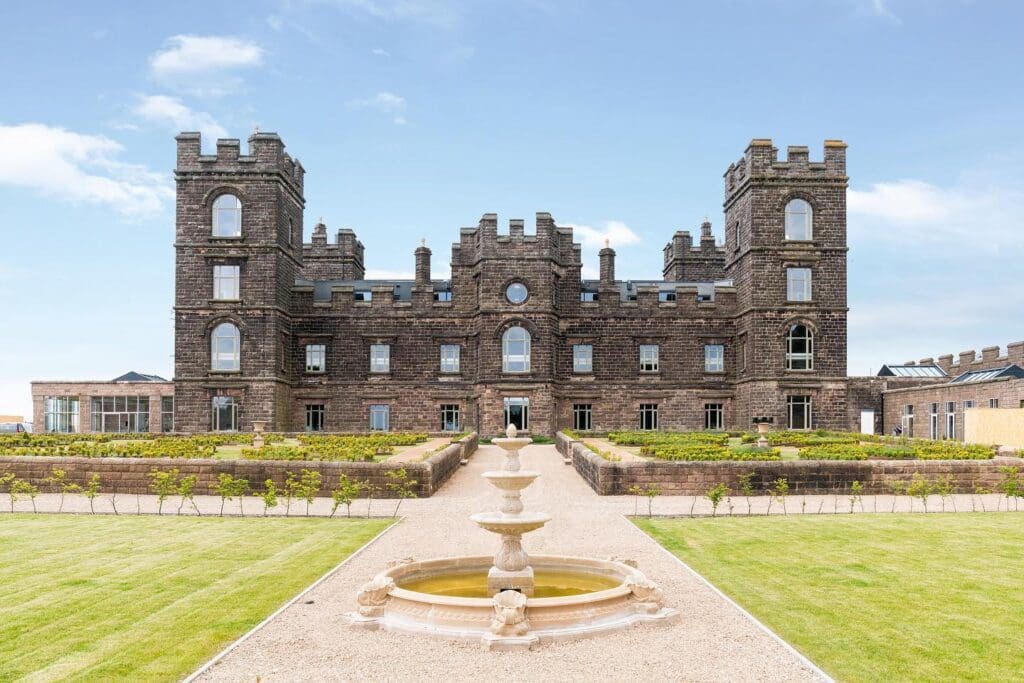 Restored exterior of Riber Castle with landscaped grounds A full-frontal view of Riber Castle in Matlock, Derbyshire, showing the dark stone façade, castellated towers, and symmetrical layout. The formal gardens feature topiary, a circular fountain in the foreground, and gravel pathways. All visible windows are custom-made timber casements from Gowercroft. Image courtesy of Dales and Peaks.