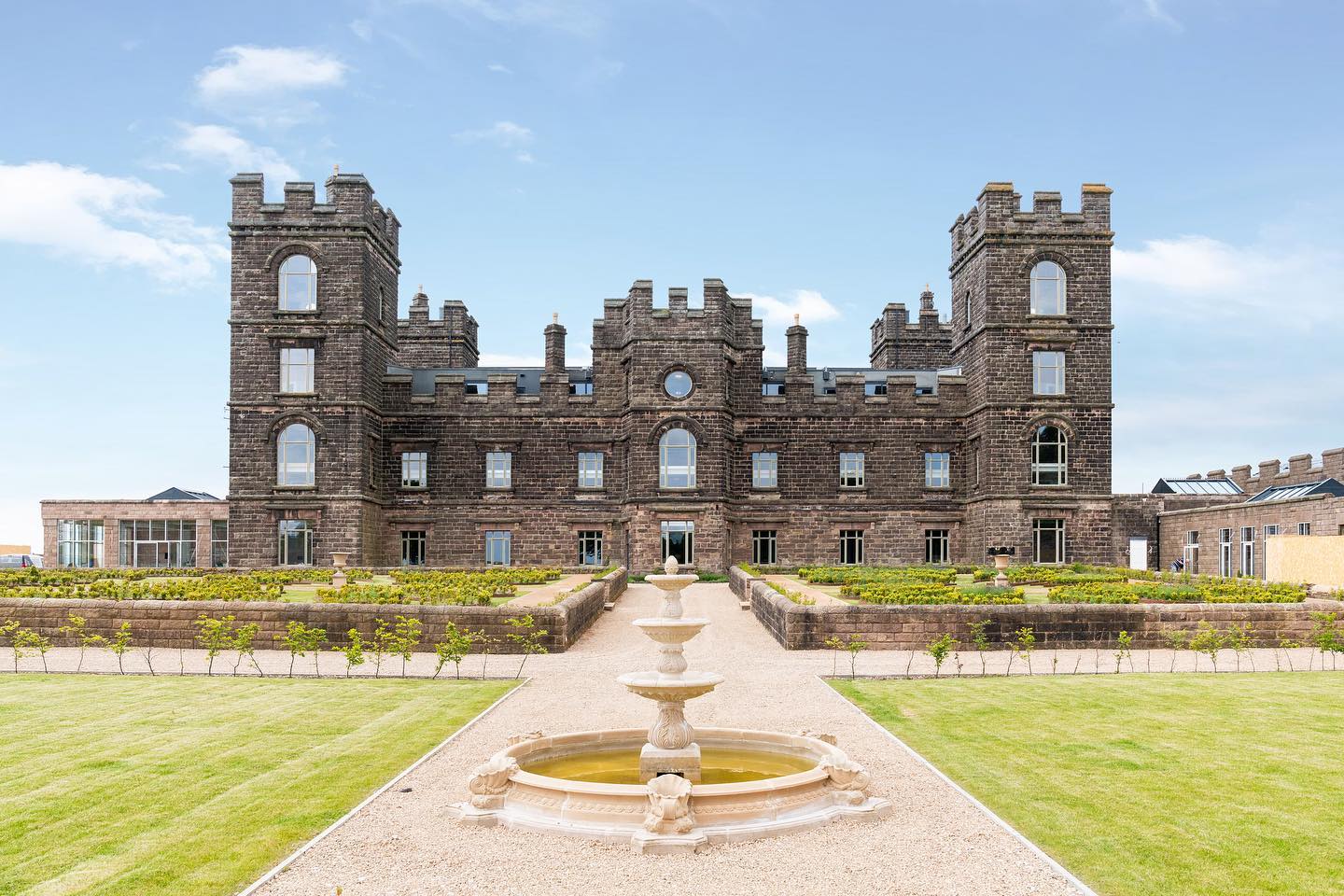 Restored exterior of Riber Castle with landscaped grounds A full-frontal view of Riber Castle in Matlock, Derbyshire, showing the dark stone façade, castellated towers, and symmetrical layout. The formal gardens feature topiary, a circular fountain in the foreground, and gravel pathways. All visible windows are custom-made timber casements from Gowercroft. Image courtesy of Dales and Peaks.