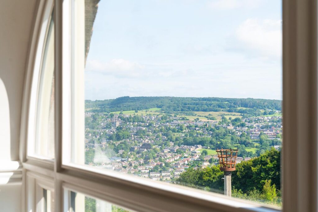 View over Matlock and the Derwent Valley from Riber Castle window
Looking out from a curved-top timber casement window at Riber Castle in Matlock, Derbyshire. The window frames a sweeping panoramic view of the town below, with rooftops, trees, and open countryside stretching into the distance. A metal beacon stands in the foreground. Image courtesy of Dales and Peaks.
