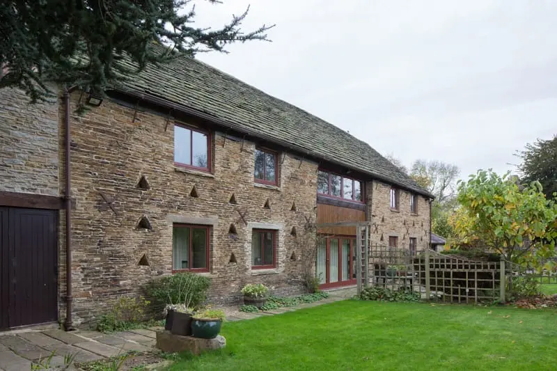 Exterior view of West Barn, a converted historic barn near Chesterfield, featuring traditional stonework with triangular ventilation holes and a roof of weathered slate tiles. The building has contemporary-style timber casement windows and doors in a muted red finish, blending modern functionality with the original rustic character. A well-kept garden and trellised entrance area sit in the foreground.