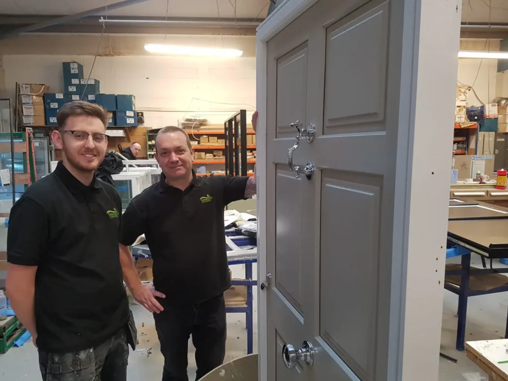 Two Gowercroft Joinery team members in the workshop, standing beside a handcrafted timber door finished in grey with polished chrome ironmongery, as part of the company’s apprentice training and development programme.