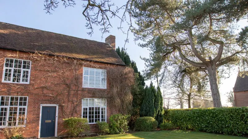 A traditional red brick farmhouse with white-painted timber windows featuring multiple panes and a dark blue front door. The house is partially covered in bare winter vines, and framed by tall trees and neatly trimmed hedges in a well-kept garden.