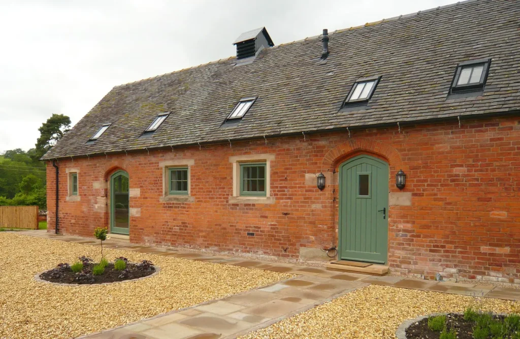 Front elevation of a brick barn conversion at Elms Farm in Snelston, Derbyshire, featuring bespoke timber casement windows and arched wooden doors painted in green, installed by Gowercroft Joinery.