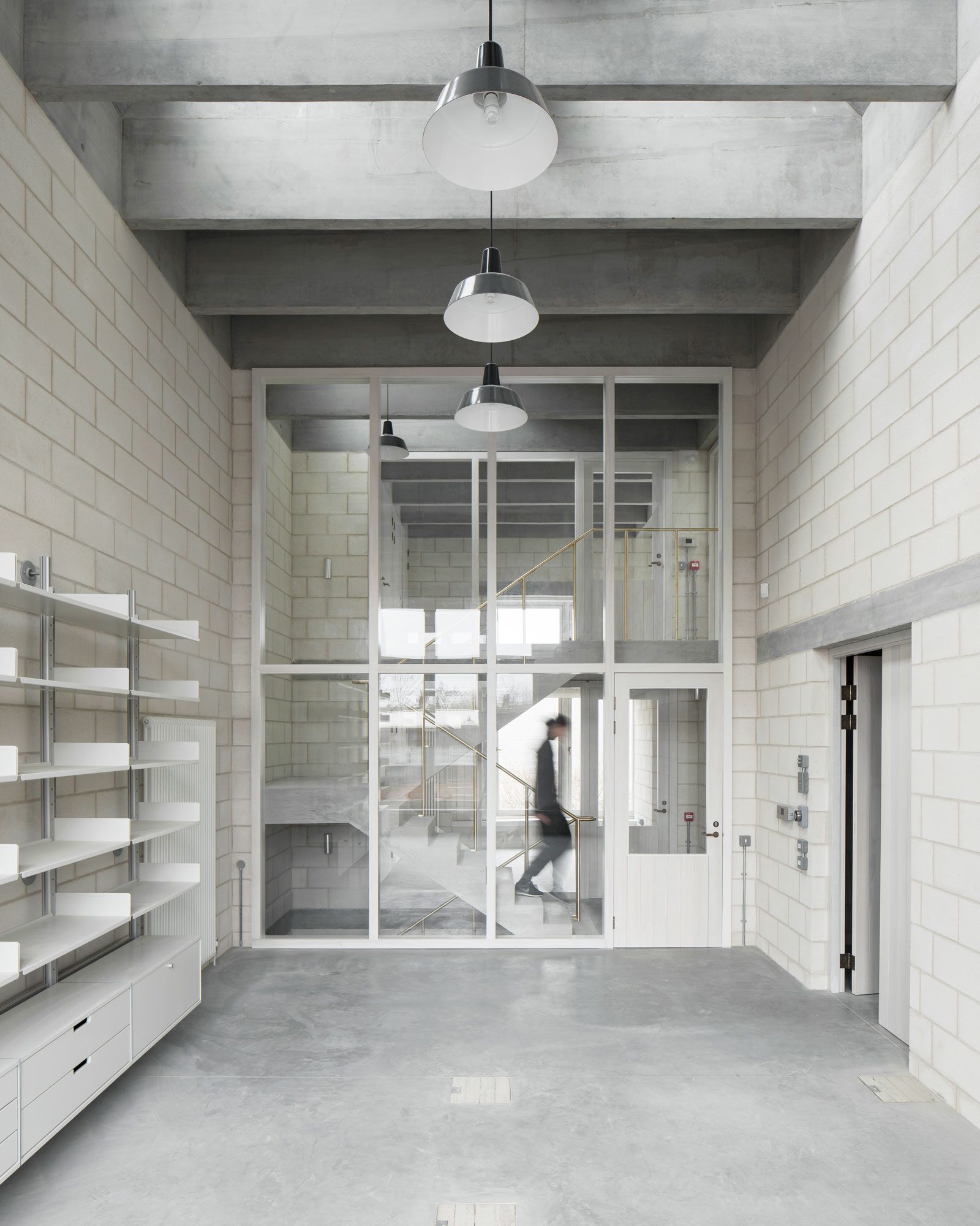 Interior view of Juergen Teller’s photography studio, showing a double-height concrete space with exposed beams and pale block walls. A large glazed timber screen frames a staircase and walkway behind, while minimalist shelving and pendant lights emphasise the raw, architectural feel. A blurred figure walks upstairs, highlighting the scale and simplicity of the space.