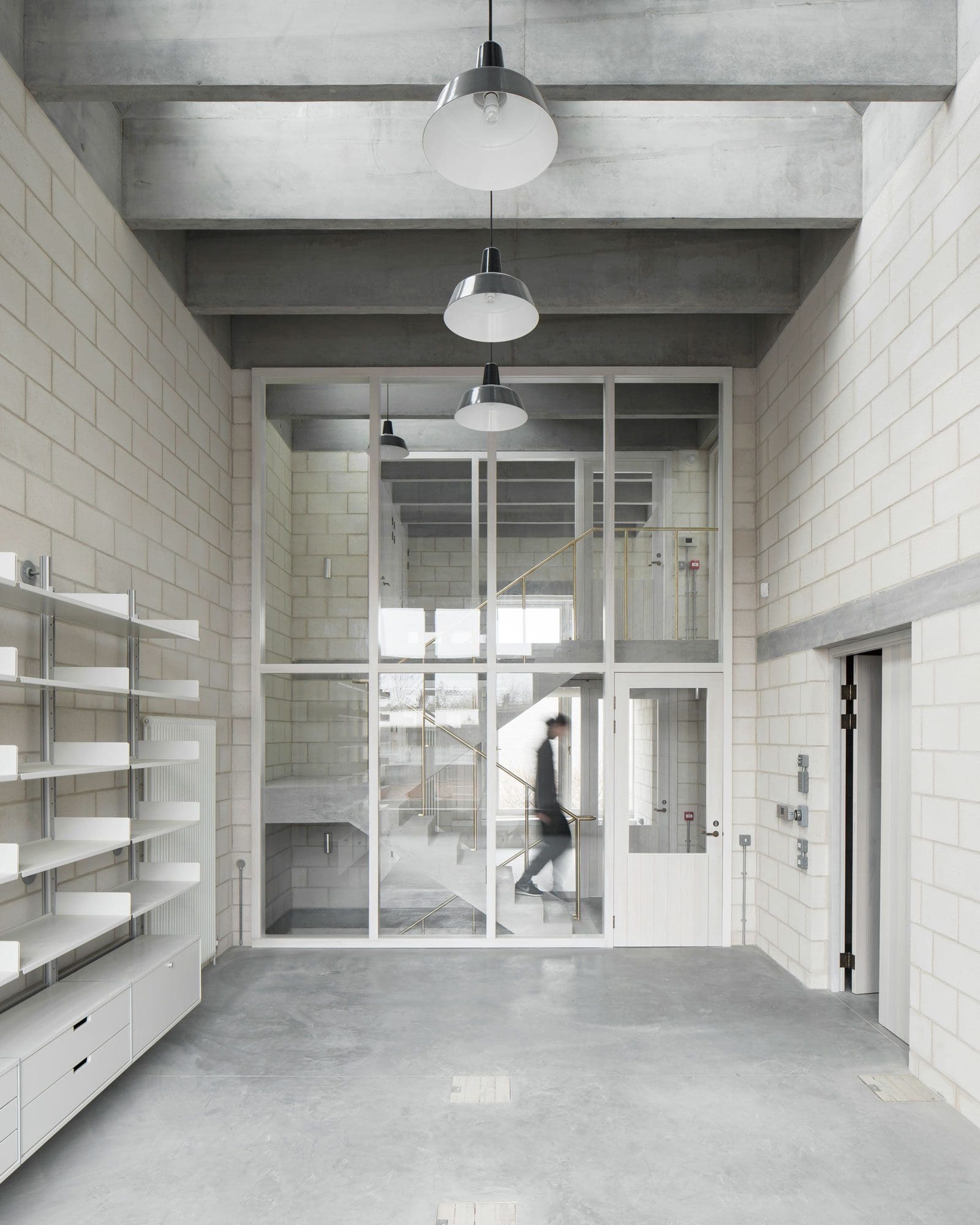 Interior view of Juergen Teller’s photography studio, showing a double-height concrete space with exposed beams and pale block walls. A large glazed timber screen frames a staircase and walkway behind, while minimalist shelving and pendant lights emphasise the raw, architectural feel. A blurred figure walks upstairs, highlighting the scale and simplicity of the space.