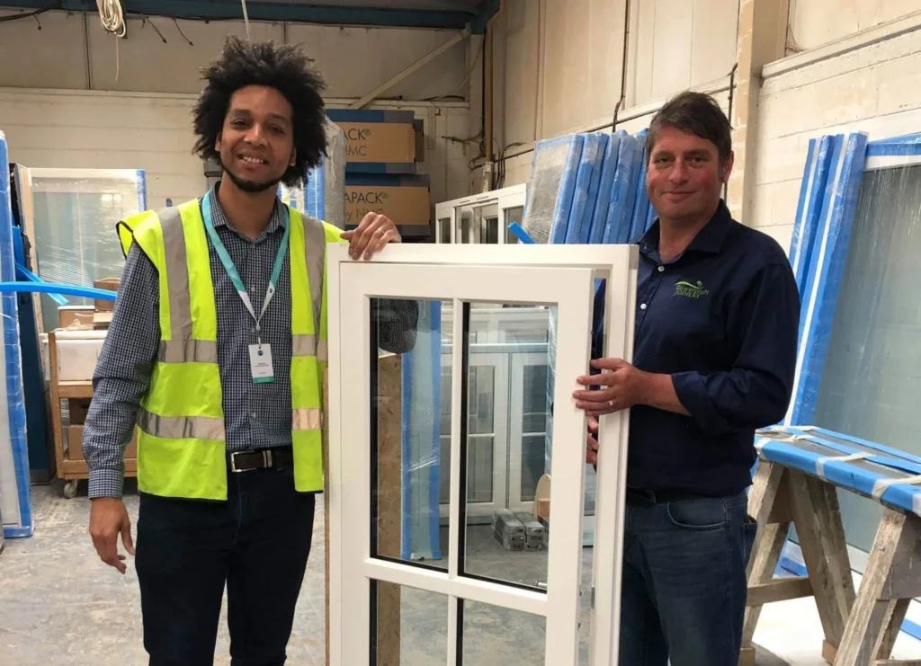 Tow men stand either side of a window at a testing centre. One wears h--vis, indicating that he is the tester and another wears a Gowercroft Joinery shirt. Both are smiling.
