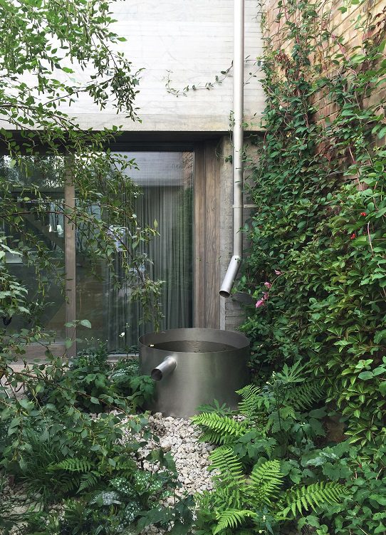 Timber-framed flush casement window by Gowercroft, set within a lush courtyard at Juergen Teller’s studio. Surrounded by climbing greenery and ferns, the window blends seamlessly into the minimalist concrete and timber palette. A stainless steel water feature and soft landscaping complete the tranquil setting.