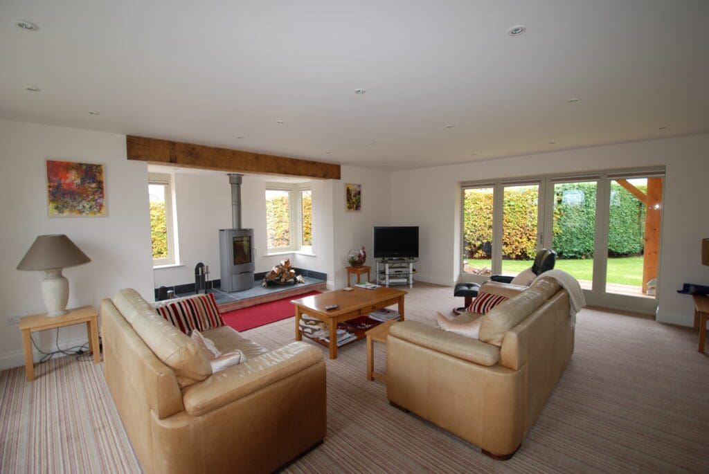 Open-plan lounge with striped carpet, leather sofas, and a wood-burning stove, surrounded by Hardwick casement windows and Tutbury bifold doors that let in plenty of natural light and offer views onto the neatly landscaped garden.