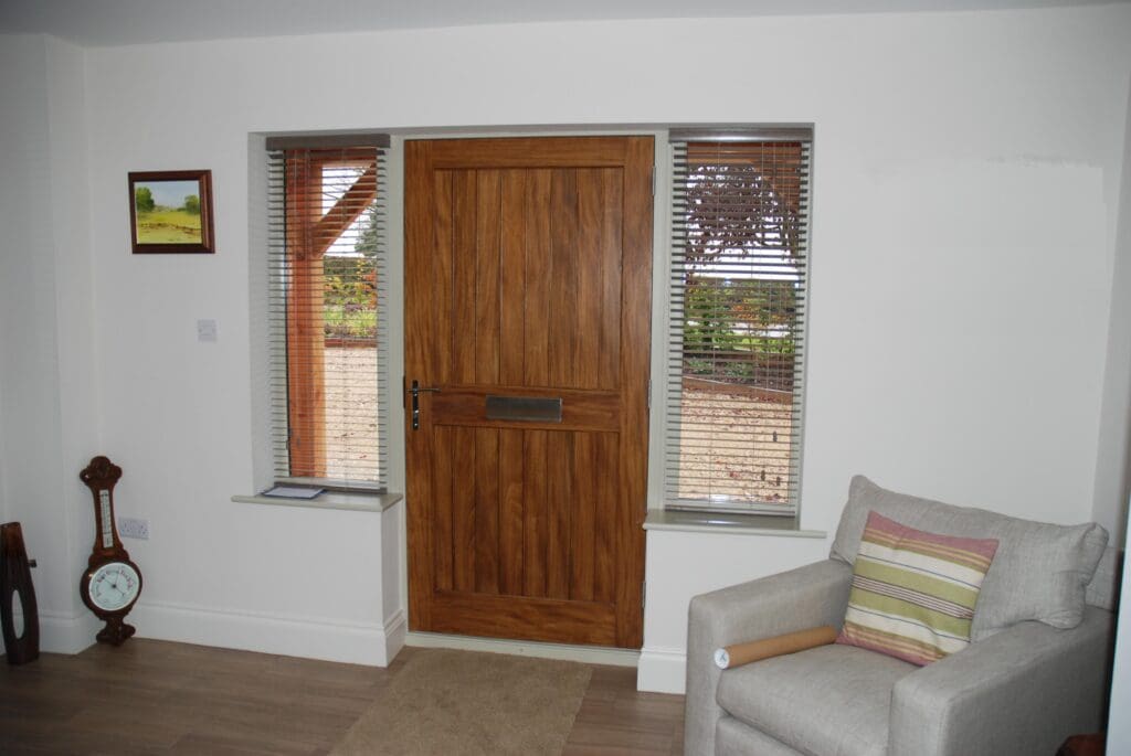 Cosy interior hallway showing a Melbourne single front door flanked by Hardwick casement winglights. All timber elements are oak stained in Walnut, and a neutral armchair sits beside the door with Venetian blinds on each glazed section.