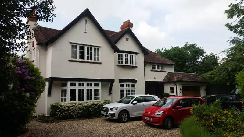 White 1930s detached home in Gerrards Cross with Georgian-style Accoya® flush casement windows and matching front and French doors, installed by Gowercroft Joinery. Three cars are parked on the gravel driveway in front of the house, surrounded by trees and shrubs.