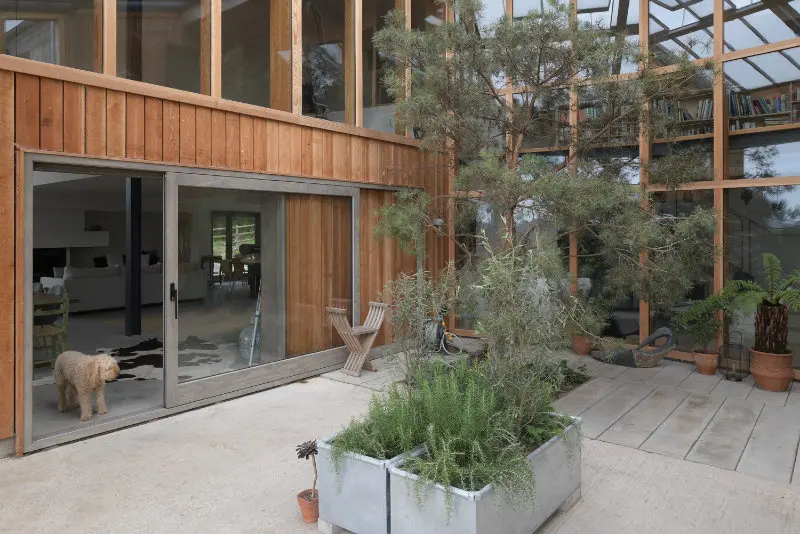 Contemporary timber-clad home in Dorset with large timber sliding doors and full-height glazed screens by Gowercroft Joinery, opening onto a modern courtyard with trees and planters. A dog stands in the doorway, emphasising homeliness.