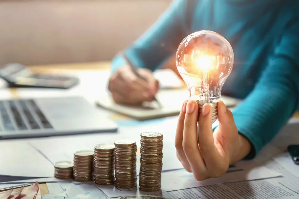Person holding a glowing light bulb next to stacked coins, with documents, a laptop, and calculator on the desk, symbolising energy savings from eco-friendly window installations.