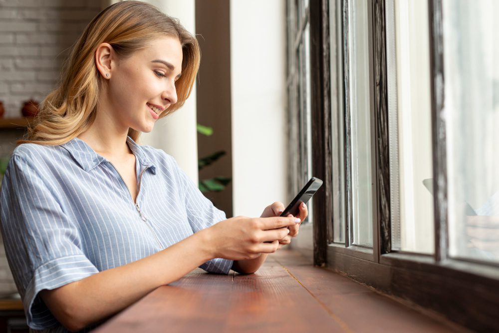 A young woman smiling and using her phone while standing beside a traditional timber sash window, with natural daylight illuminating her face.