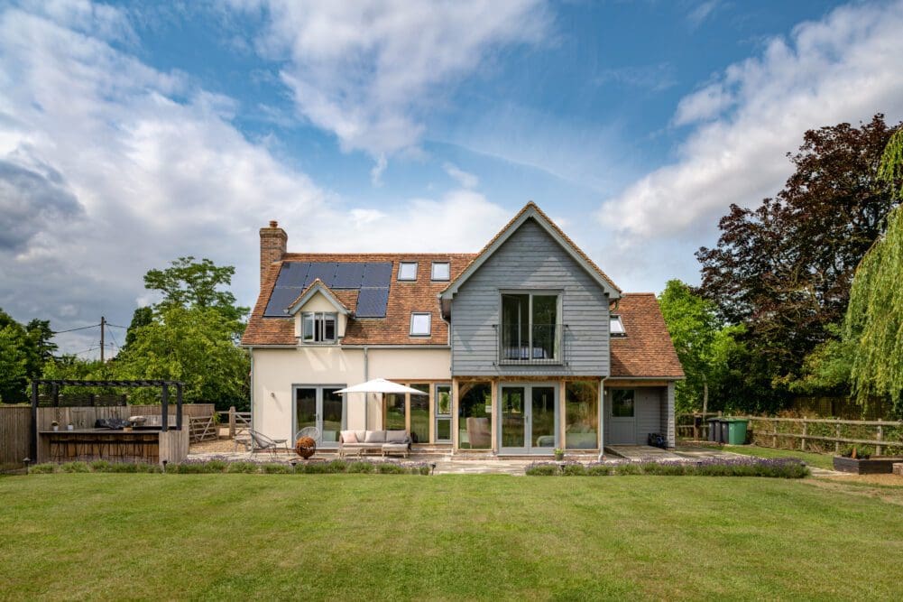 The back of a welcoming two-story home showcasing solar panels on the roof, a gray weatherboard extension, and large glass doors that open up to a well-manicured lawn with an outdoor seating area under a clear sky.