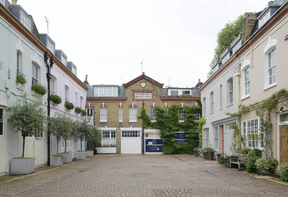A charming cobbled mews street lined with pastel-coloured and white-painted historic buildings, featuring sash windows and greenery in planters. At the far end, a larger period property displays a plaque reading "Horbury Mews 1878", indicating its heritage status. This picturesque scene highlights the type of traditional architecture where energy-efficient windows may need to be sensitively integrated.