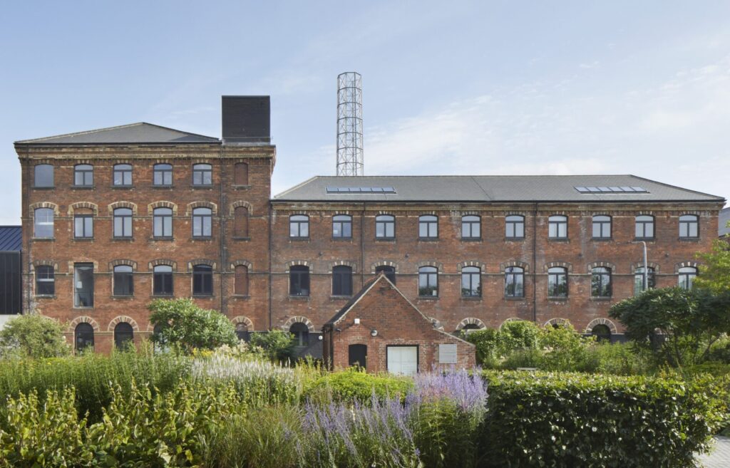 The image depicts a regenerated mill building with a historic red brick facade, showcasing its industrial heritage. The building has 219 windows, all from Gowercroft's Timber Hardwick casement range, which seamlessly integrate contemporary functionality with traditional aesthetics. The red bricks are interspersed with lighter brick detailing around the windows and edges, adding texture and visual interest. The windows are uniformly arranged across the facade. The building features modern architectural elements, including a sleek black extension and a tall, lattice-style chimney. The surrounding area is landscaped with lush greenery and flowering plants, adding a touch of nature to the urban environment. The sky is clear and blue, highlighting the clean lines and structural details of the building.