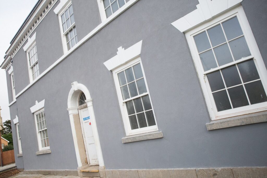 A close-up view of a period-style building with traditional sash windows, featuring white window frames and decorative bars dividing the glass panes. The architectural detail showcases a clear example of astragal bars, ideal for visually distinguishing between authentic glazing bars and plant-on alternatives - relevant for understanding window style choices in heritage or conservation settings.
