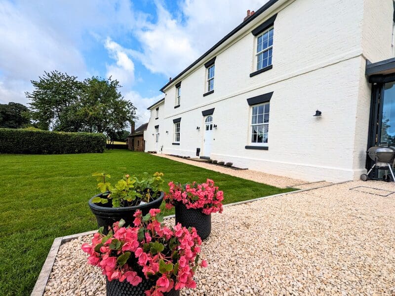 A beautifully renovated white brick house with well-maintained green lawn and blooming flower pots in the foreground. The house features Gowercroft Joinery's timber windows, including Winston Heritage Sliding Sash Windows and a Churchill Heritage Door. The windows and door seamlessly blend with the historical aesthetic of the property, showcasing a successful combination of heritage design and modern functionality.