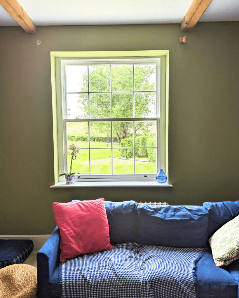 An interior view of a traditional sash-style heritage window with elegant timber framing, set in an olive green wall above a cosy navy-blue sofa. The window offers a clear view of a lush garden and mature tree, illustrating how Gowercroft Joinery’s heritage windows preserve historical character while enhancing energy efficiency and comfort in period homes.