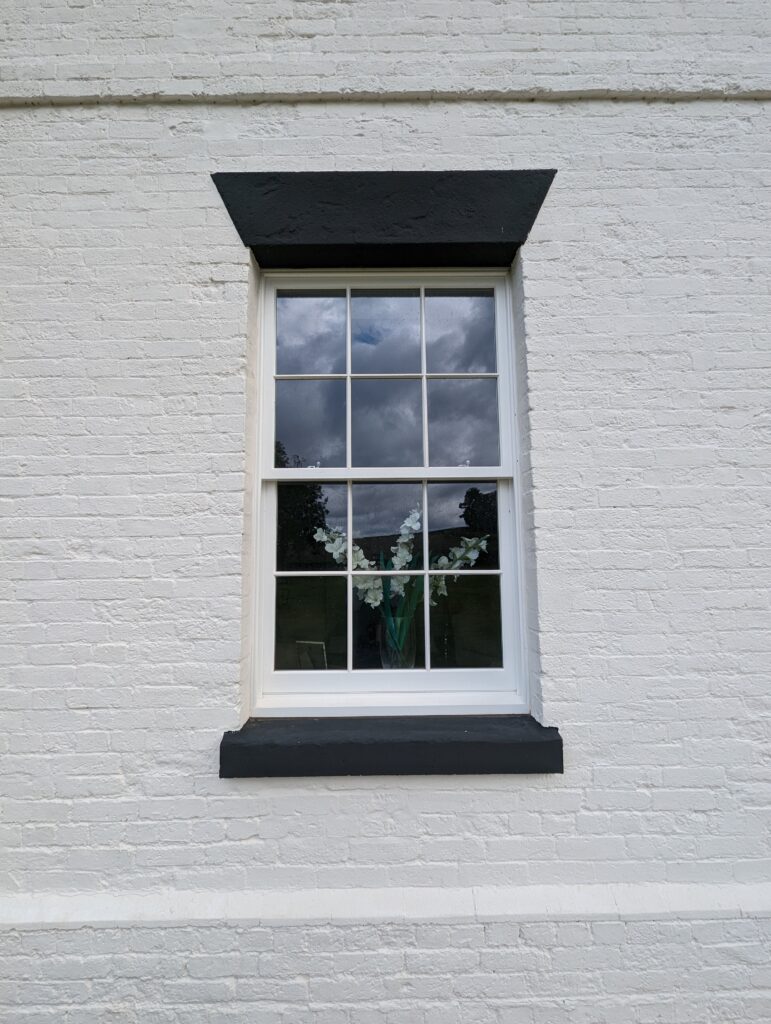 A close-up of a white brick wall with a single sash window featuring integrated conservation bars. The window is framed with a black lintel and sill, contrasting sharply with the white wall. Through the window, a vase with white flowers is visible, and the reflection of a cloudy sky can be seen in the glass. The window's traditional design enhances the classic aesthetic of the building's exterior