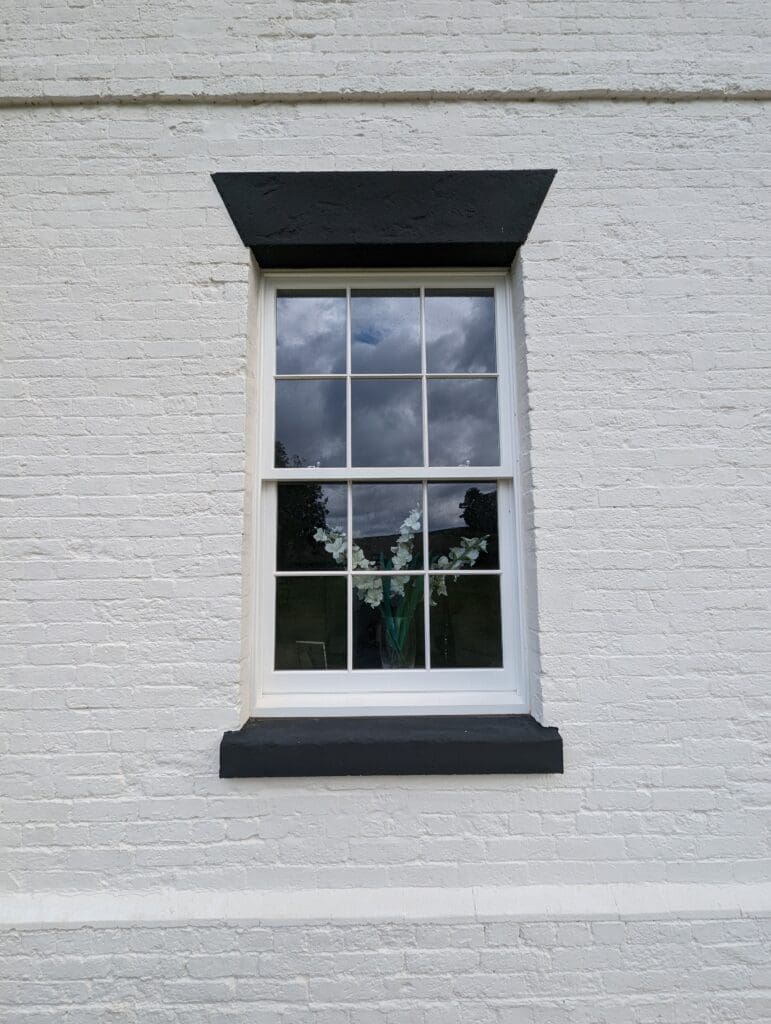 A close-up of a white brick wall with a single sash window featuring integrated conservation bars. The window is framed with a black lintel and sill, contrasting sharply with the white wall. Through the window, a vase with white flowers is visible, and the reflection of a cloudy sky can be seen in the glass. The window's traditional design enhances the classic aesthetic of the building's exterior