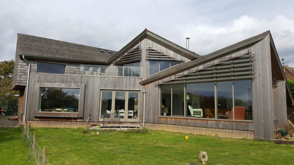 A striking modern home with sharp angular rooflines and extensive timber cladding, featuring large glass windows and clean architectural lines. The exterior showcases the natural weathering of Accoya wood, known for its durability, stability, and sustainable credentials.