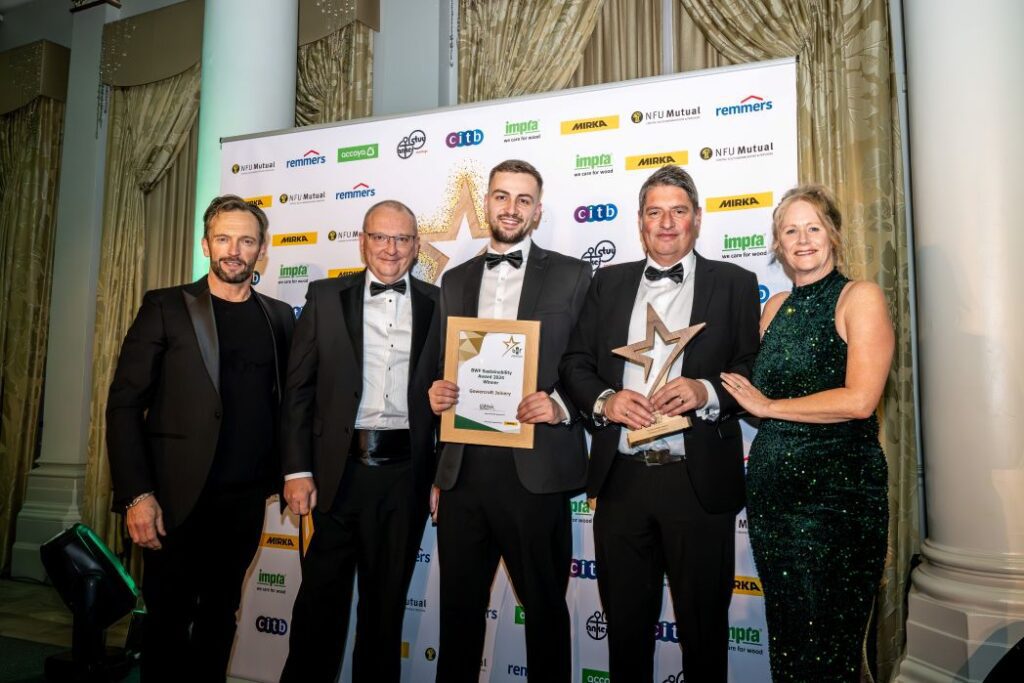 Group photo at the BWF Sustainability Awards 2024 featuring Harry Dixon, Finance Director of Gowercroft Joinery, holding the Sustainability Award certificate, and Darren Young, Production Director, holding the trophy. They are joined by the event host, a judge, and Helen Hewitt, Chief Executive of the British Woodworking Federation, on the right. All are dressed in formal evening wear, standing in front of a branded sponsor wall with elegant curtains and pillars in the background.