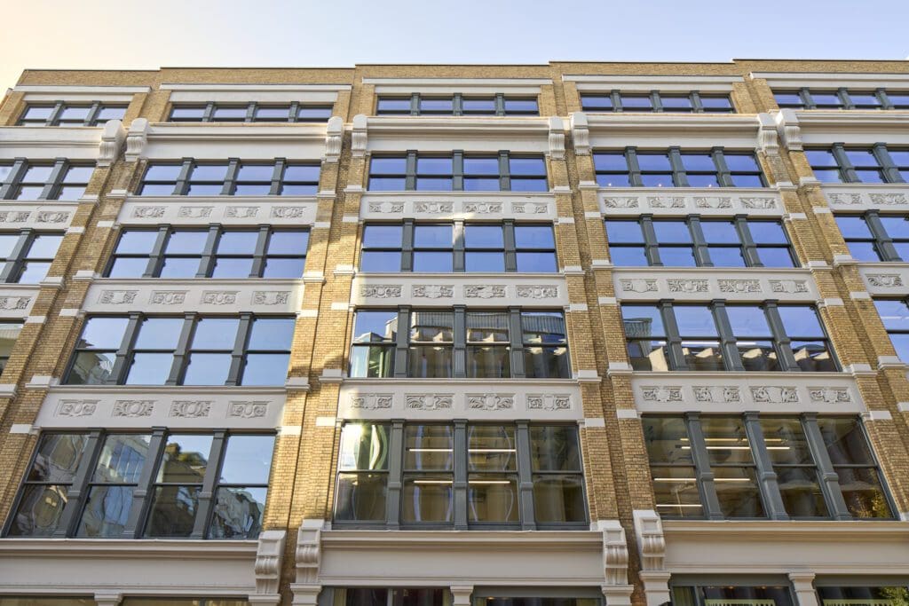 Front elevation of The Waterman Building, Farringdon, London – A close-up of the restored front façade, showing rows of traditional-style sliding sash windows framed by intricate white stonework. The black window frames contrast against the golden brick exterior, preserving the heritage character of this historic London warehouse.