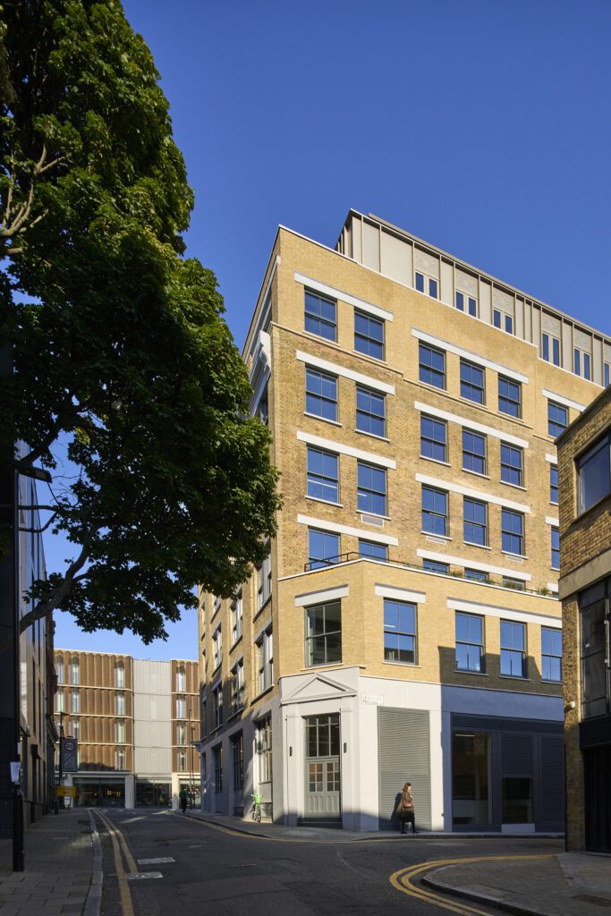 Corner perspective of The Waterman Building, Islington, London – A street-level view of the corner of this Grade II listed warehouse, where a modern rooftop extension blends with the traditional brick and stone detailing below. Large sash windows reflect the blue sky, while a tree-lined street adds greenery to the urban surroundings.