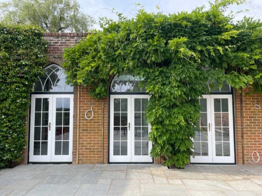A row of elegant heritage-style French doors with arched fanlights above, set in a traditional red-brick building partly covered in lush green climbing plants. The crisp white timber frames and clear glazing exemplify how modern vacuum glazing technology can be seamlessly integrated into classic architectural designs, marking a decade of innovation in enhancing energy efficiency without compromising heritage aesthetics.
