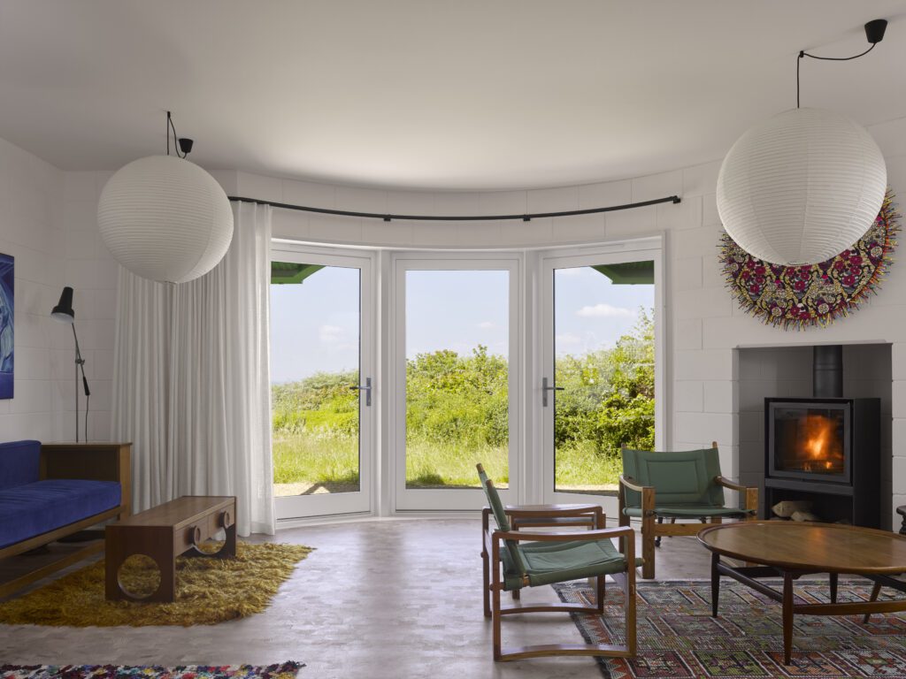 Curved Melbourne French doors: This image shows a bright, airy living room with white Melbourne timber French doors on the interior, featuring a curved design that opens up to the lush Dorset countryside. The doors, framed in crisp white, allow plenty of natural light to fill the room, while the curved structure creates a seamless transition between the indoors and outdoors. The room features light-toned furnishings, including a blue sofa and wooden coffee table, paired with large round paper lanterns and a modern fireplace. The soft, warm light enhances the peaceful atmosphere, drawing attention to the minimalist yet inviting design