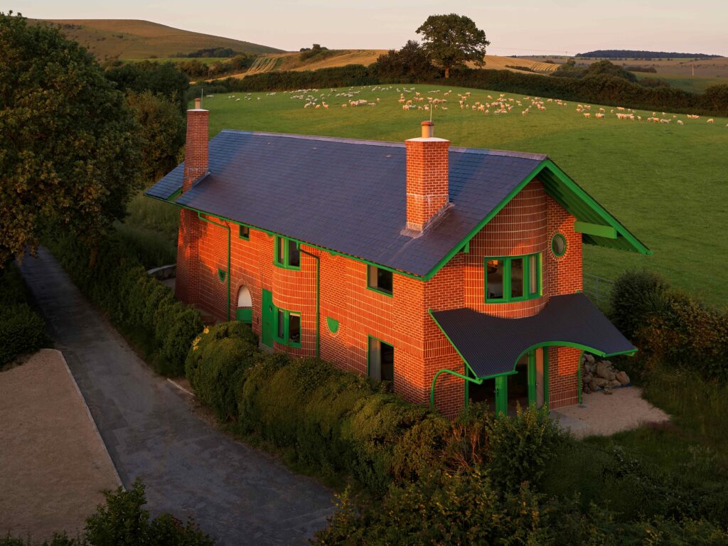 Red House External Elevation with Hardwick Casements and Melbourne Doors: An exterior shot of The Red House, showcasing the building's bold design. The handmade red brickwork contrasts with bright green Hardwick casement windows and Melbourne timber doors, which punctuate the façade. The house, sitting on a hillside amidst rolling fields, features exaggerated overhanging green eaves and a steep-pitched slate roof. The vibrant green timber windows and doors add a playful touch to the traditional brick structure, with the countryside extending into the distance, reinforcing the house's connection with its natural surroundings.