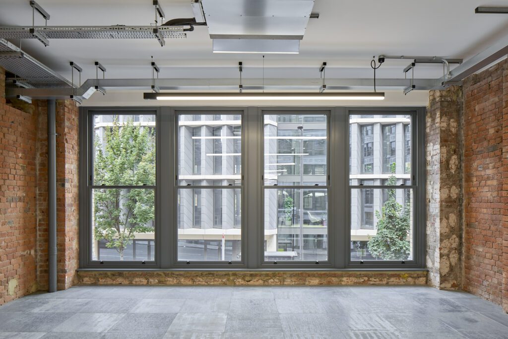 A set of four large Chatsworth sliding sash windows in The Waterman Building, Farringdon, London, framed by exposed brick walls. The windows, featuring slim timber profiles in a muted grey finish, allow natural light to flood the contemporary office space. The irregular sizes reflect the historic character of the warehouse conversion, offering energy efficiency while preserving the industrial charm. Through the glass, a modern London office building with sleek architectural lines contrasts against the heritage interior.