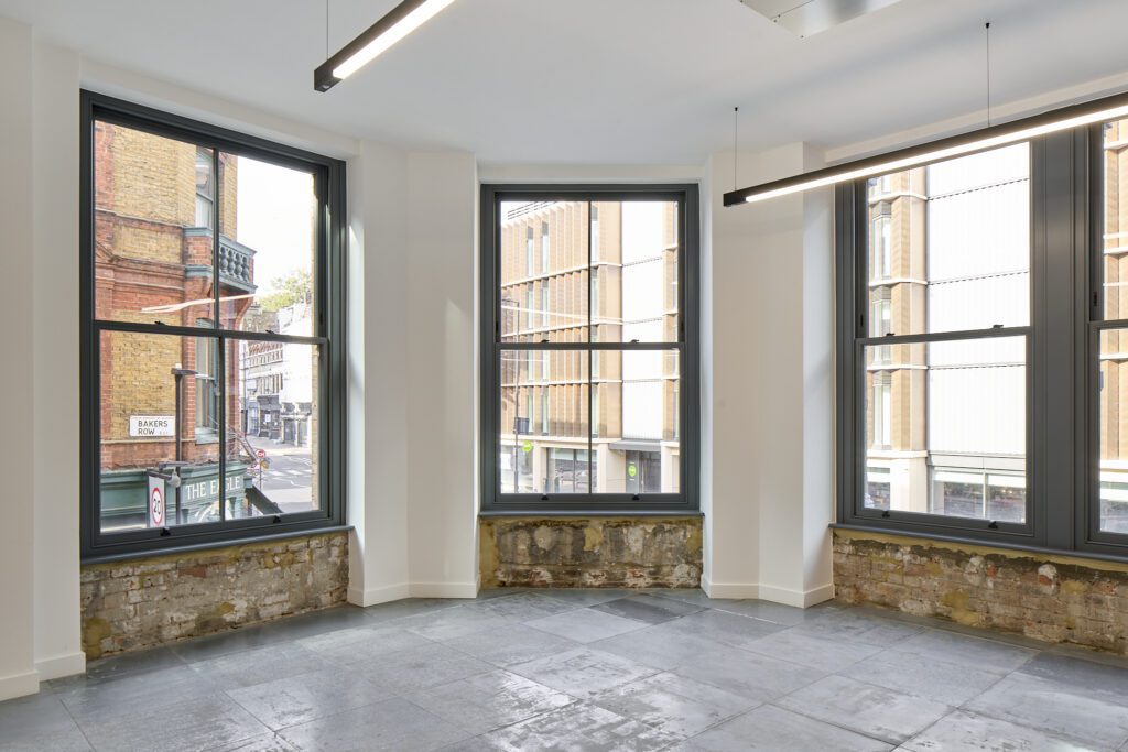A bright corner space in The Waterman Building, Farringdon, London, featuring large Chatsworth sliding sash windows. The timber-framed windows, finished in a deep grey, allow natural light to flood the room while offering a striking contrast to the exposed brick and modern interior. The varied window heights reflect the historic character of the conservation area. Through the glass, a view of the bustling London streets, including Bakers Row and The Eagle pub, connects the heritage-rich interior with the city’s architectural blend of old and new.
