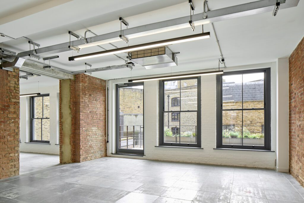 Interior view of The Waterman Building, Farringdon, London – A long, open-plan office space featuring industrial-style lighting, exposed ceiling ductwork, and a row of large timber sash windows of varying heights. The windows provide views over the surrounding cityscape while allowing natural light to flood the room.