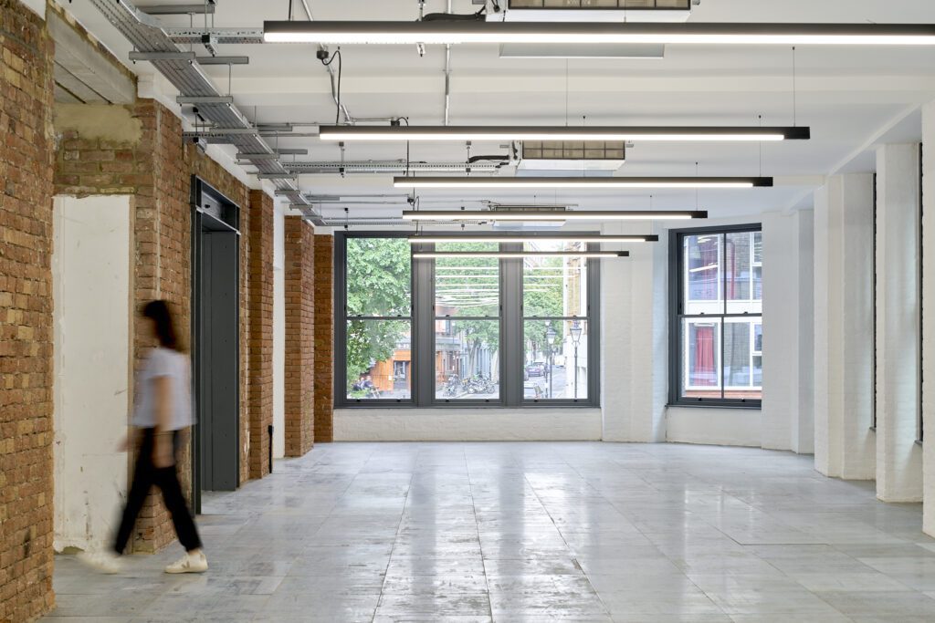 Long open-plan office with heritage sash windows and exposed brick – The Waterman Building, Farringdon, London
A spacious open-plan office featuring exposed brick walls, polished concrete floors, and large Chatsworth sliding sash windows that allow natural light to flood the space. The industrial ceiling showcases metal beams and modern linear lighting. On the left, an elevator is inset into the brick wall, blending seamlessly into the building’s restored heritage features.