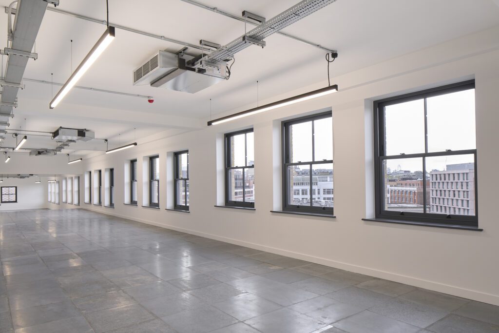 A spacious, modern office interior in The Waterman Building, Farringdon, London, featuring a row of tall, grey-framed Chatsworth sliding sash windows. The windows vary in height and size, reflecting the irregular dimensions of the original historic warehouse structure. The polished concrete floor and exposed industrial-style ceiling fixtures contrast with the restored heritage windows, offering a bright, energy-efficient workspace with panoramic views of the London skyline.