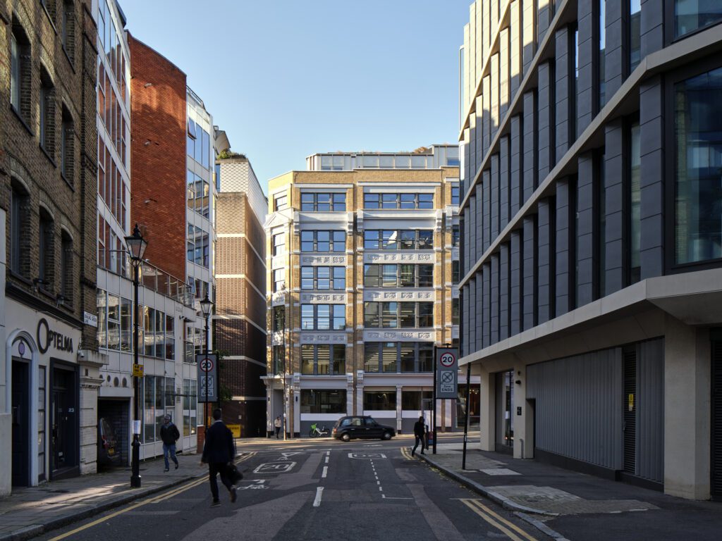 A view down a quiet street in Farringdon, London, leading to The Waterman Building, a beautifully restored Victorian warehouse featuring Gowercroft's Chatsworth sliding sash windows. The façade showcases elegant white detailing against the original brickwork, seamlessly integrating heritage aesthetics with modern energy efficiency. The surrounding urban environment blends historic and contemporary architecture, highlighting the building’s transformation into a sustainable, low-carbon office space. Pedestrians and cyclists navigate the area, reinforcing its role as a vibrant part of the London Borough of Islington.