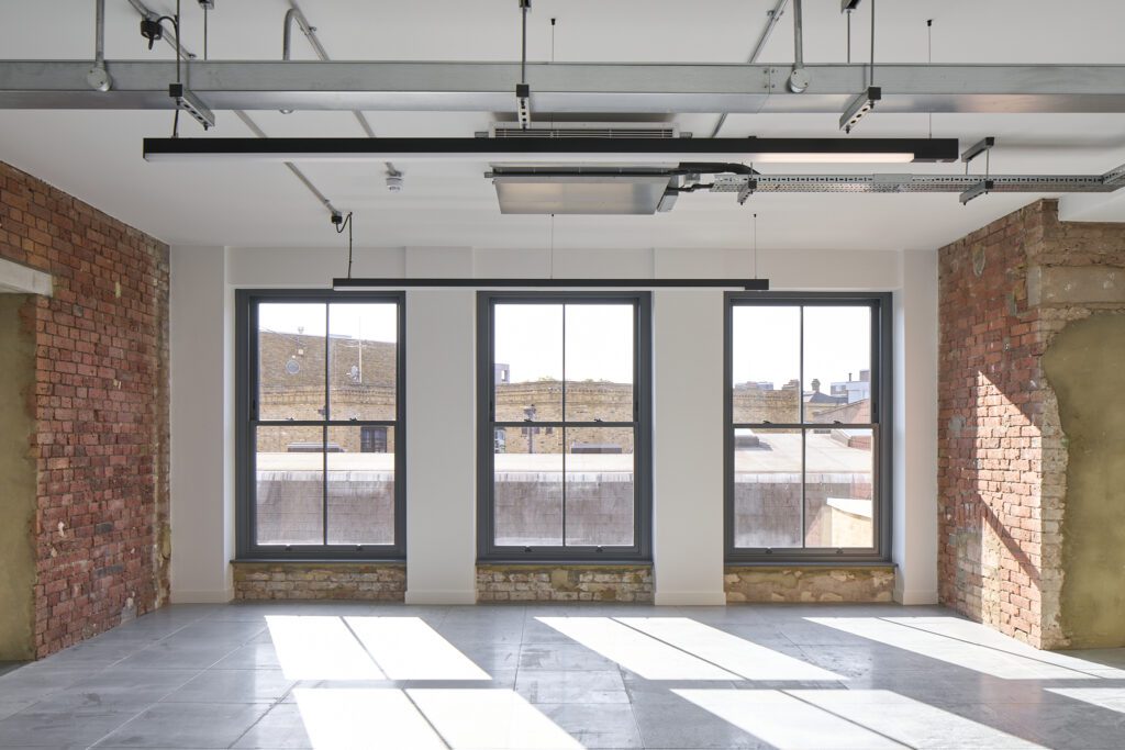 Renovated workspace inside The Waterman Building, Farringdon, London – A bright, open-plan office space featuring exposed brick walls, polished concrete floors, and large sash windows that let in streams of natural light. The windows are set into deep reveals, emphasising the building’s solid historic construction.