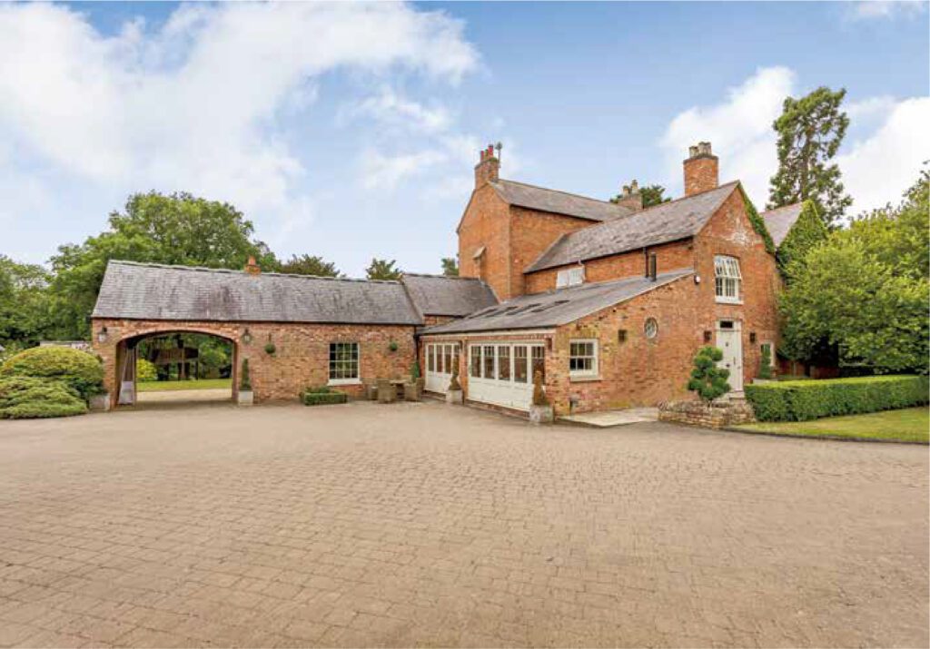 A beautifully restored traditional brick country house with a mix of pitched and flat roofs. The property features elegant timber sliding sash windows and a Churchill-style entrance door, blending heritage charm with modern craftsmanship. A large paved driveway leads up to the house, surrounded by neatly trimmed hedges and lush greenery.