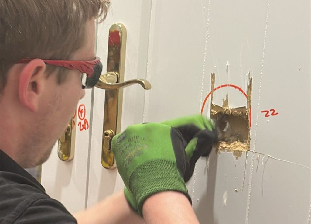 Technician performing PAS 24 security test by attempting forced entry near the lock of a timber door using hand tools.